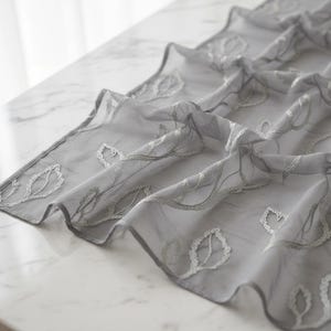 Può includere: Un runner da tavolo grigio trasparente con ricami bianchi a forma di foglie e tralci. Il tessuto è drappeggiato su una superficie, mettendo in mostra il design e la trama delicati. Il runner aggiunge un tocco elegante.