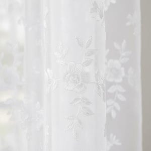 May include: Close-up of a sheer white curtain with embroidered floral and leaf patterns. The delicate design features detailed white flowers and foliage, creating a soft, textured look. The curtain is semi-transparent, allowing light to filter through.