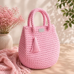 Può includere: Una borsa a mano all'uncinetto rosa chiaro con una silhouette arrotondata, due robusti manici intrecciati e un ciondolo a nappa rosa abbinato attaccato a un manico. Una piccola piastra metallica rettangolare argentata vuota è centrata sul davanti.