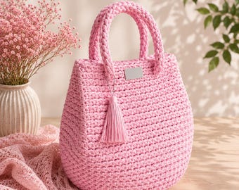 May include: A light pink crochet handbag featuring a rounded silhouette, two sturdy braided handles, and a matching pink tassel charm attached to one handle. A small, rectangular, blank silver-toned metal plate is centered on the front exterior.