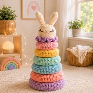 May include: A crochet stacking toy featuring a cream-colored rabbit head topper with embroidered eyes, followed by a ruffled purple collar and six stacked rings in pastel shades of pink, yellow, mint green, light blue, peach, and lavender.