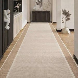 May include: A hallway featuring a beige rug with a light brown border. Dark cabinets with glass doors and a white sculpture are on the left. A dark brown cabinet with artwork and plants is at the end.