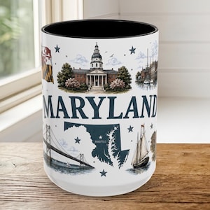 May include: A white ceramic mug with a black interior featuring a Maryland state theme. The design includes the Maryland state flag, the state capitol building, a map of Maryland with Annapolis labeled, a suspension bridge, and a sailboat.