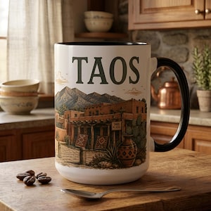 May include: A white ceramic mug with a black interior and handle, featuring a printed illustration of Taos Plaza with mountains, adobe buildings, a decorative vase, and the word TAOS in large green letters. The mug sits on a wooden surface.