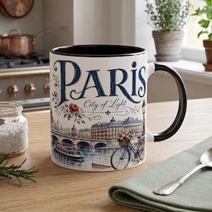 May include: A white ceramic mug with a black interior and handle, featuring a blue and multi-colored illustration of a Parisian river scene. The text PARIS City of Light is printed in large blue script above a bridge, a boat, and a bicycle.
