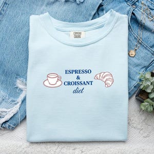 Può includere: Una t-shirt azzurro chiaro con un design grafico che mostra una piccola tazzina da caffè su un piattino, un croissant e il testo blu scuro "ESPRESSO & CROISSANT diet" stampato al centro. La maglietta ha un'etichetta del marchio Comfort Colors.