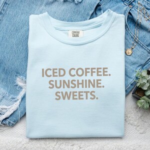 Könnte beinhalten: Ein hellblaues T-Shirt mit dem Aufdruck "ICED COFFEE. SUNSHINE. SWEETS." in Braun. Das Shirt liegt gefaltet auf einer blauen Jeans. Eine kleine Sukkulente und Schmuck sind ebenfalls zu sehen.