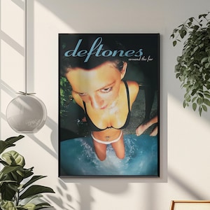 May include: A framed poster of the band Deftones, with the text "deftones around the fur" in blue. The image shows a person in a black bikini in water, viewed from above. The poster is displayed on a white wall.