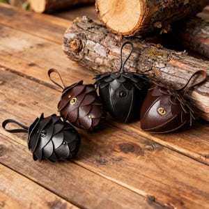 May include: Four small pouches shaped like acorns or pinecones, crafted from black and dark brown leather. Each pouch features overlapping petal-like layers, a top loop for hanging, and a central metallic twist-lock closure on the front.