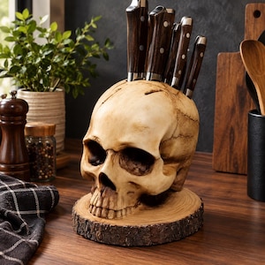 May include: A skull-shaped knife block made of off-white resin, featuring five slots holding kitchen knives with dark brown wooden handles. The skull rests on a circular base made of a natural wood slice with visible bark edges and grain.