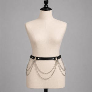 May include: A black leather belt featuring two silver-toned metal rings on the front. Multiple draped silver-toned metal chains hang from the rings, creating a layered effect across the front of the belt. The belt is secured with a metal buckle.