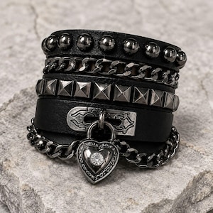 May include: A black multi-layered cuff bracelet featuring a row of round studs, a metallic chain, a row of pyramid studs, and a central strap with a decorative silver-toned plate. A heart-shaped padlock charm with a clear crystal center hangs below.
