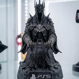 Puede incluir: Estatua negra de un personaje de El Señor de los Anillos, sentado con corona y armadura. La estatua está sobre una base rocosa con el logotipo de PlayStation 5.