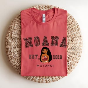 May include: A coral-coloured t-shirt with the word "MOANA" in a collegiate font. Below the word is an illustration of a character and the text "EST. 2016" and "MOTUNUI". The shirt is displayed on a woven mat.