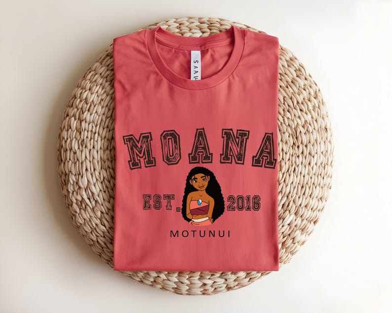 May include: A coral-coloured t-shirt with the word "MOANA" in a collegiate font. Below the word is an illustration of a character and the text "EST. 2016" and "MOTUNUI". The shirt is displayed on a woven mat.