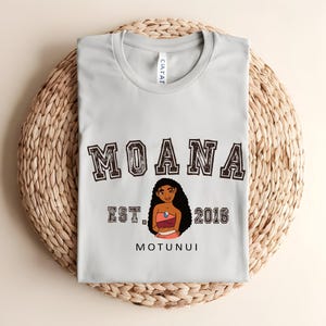 May include: A pale grey t-shirt with the word "MOANA" in a collegiate font above a character illustration. Below the illustration are the words "EST. 2016" and "MOTUNUI". The shirt is folded on a woven, circular surface.