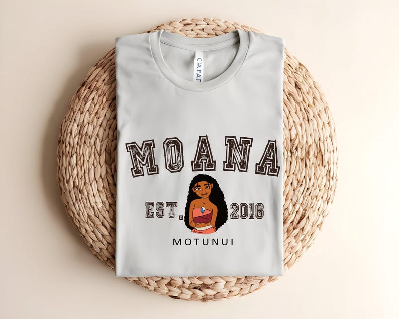 May include: A pale grey t-shirt with the word "MOANA" in a collegiate font above a character illustration. Below the illustration are the words "EST. 2016" and "MOTUNUI". The shirt is folded on a woven, circular surface.