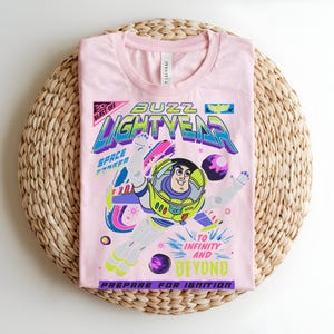May include: A pale pink t-shirt with a graphic of Buzz Lightyear. The design includes the words "Buzz Lightyear" and "To Infinity and Beyond." The shirt is folded on a woven, circular mat.