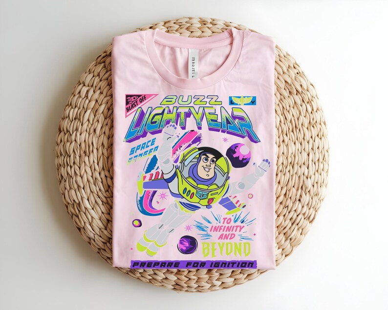 May include: A pale pink t-shirt with a graphic of Buzz Lightyear. The design includes the words "Buzz Lightyear" and "To Infinity and Beyond." The shirt is folded on a woven, circular mat.