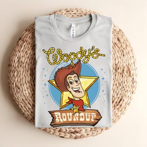 Puede incluir: Camiseta gris claro con una imagen de dibujos animados de Woody de Toy Story. El dise&ntilde;o incluye la cara de Woody dentro de una estrella dorada, un c&iacute;rculo azul y las palabras "Woody's Roundup" en una fuente de estilo occidental. La camiseta est&aacute; doblada sobre una superficie tejida.