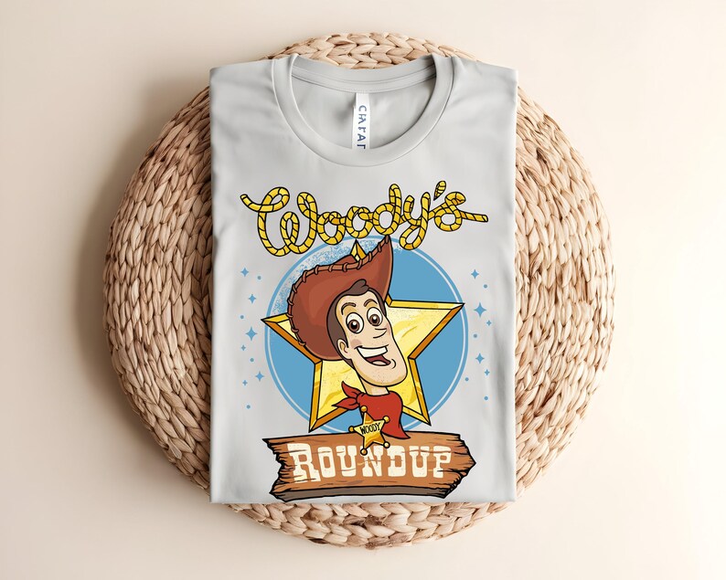 Puede incluir: Camiseta gris claro con una imagen de dibujos animados de Woody de Toy Story. El dise&ntilde;o incluye la cara de Woody dentro de una estrella dorada, un c&iacute;rculo azul y las palabras "Woody's Roundup" en una fuente de estilo occidental. La camiseta est&aacute; doblada sobre una superficie tejida.
