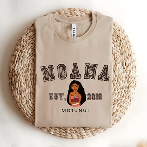 May include: Beige crew neck t-shirt featuring the word "MOANA" in a collegiate style font. Below is an illustration of a cartoon character and the text "EST. 2016 MOTUNUI". The shirt is folded on a woven, circular mat.