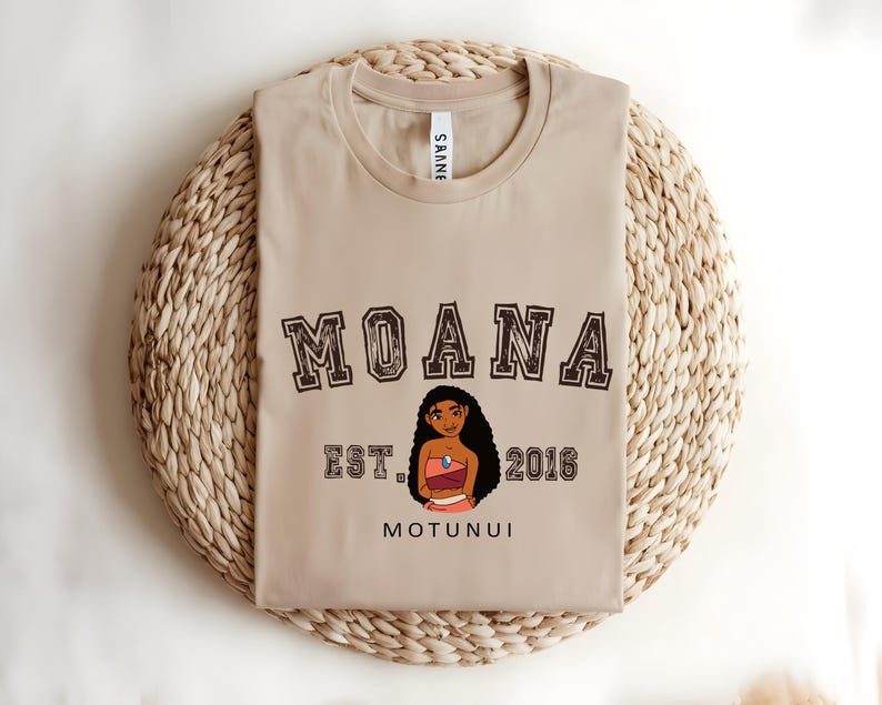 May include: Beige crew neck t-shirt featuring the word "MOANA" in a collegiate style font. Below is an illustration of a cartoon character and the text "EST. 2016 MOTUNUI". The shirt is folded on a woven, circular mat.