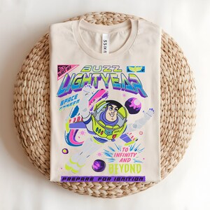 May include: Beige t-shirt with a vibrant Buzz Lightyear graphic from Toy Story. The design includes the text "Buzz Lightyear" and "To Infinity and Beyond." The shirt is folded on a woven mat.