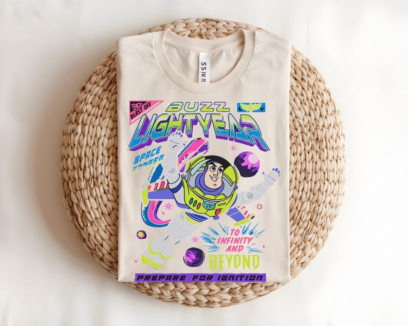 May include: Beige t-shirt with a vibrant Buzz Lightyear graphic from Toy Story. The design includes the text "Buzz Lightyear" and "To Infinity and Beyond." The shirt is folded on a woven mat.