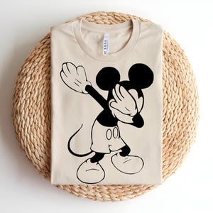 K&ouml;nnte beinhalten: Ein beiges T-Shirt mit Rundhalsausschnitt und einem schwarzen Grafikdruck der Zeichentrickfigur Mickey Mouse, die eine Dab-Pose ausf&uuml;hrt. Das Design zeigt die Figur im Profil mit gro&szlig;en Ohren, Shorts und Handschuhen, zentriert auf der Vorderseite.