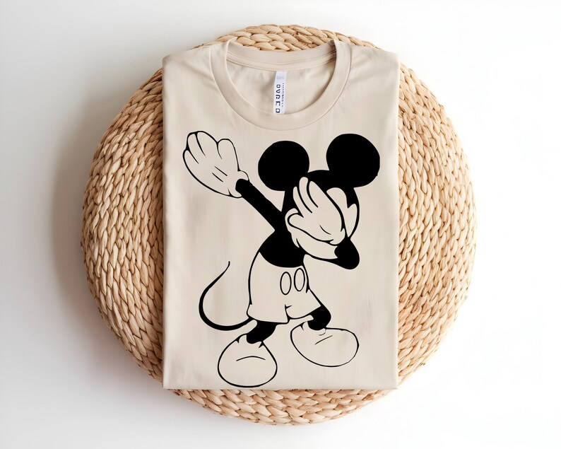 K&ouml;nnte beinhalten: Ein beiges T-Shirt mit Rundhalsausschnitt und einem schwarzen Grafikdruck der Zeichentrickfigur Mickey Mouse, die eine Dab-Pose ausf&uuml;hrt. Das Design zeigt die Figur im Profil mit gro&szlig;en Ohren, Shorts und Handschuhen, zentriert auf der Vorderseite.