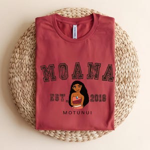May include: A maroon t-shirt with the word "MOANA" in a collegiate font above a cartoon character illustration. Below the illustration are the words "EST. 2016" and "MOTUNUI". The shirt is folded on a woven, circular surface.