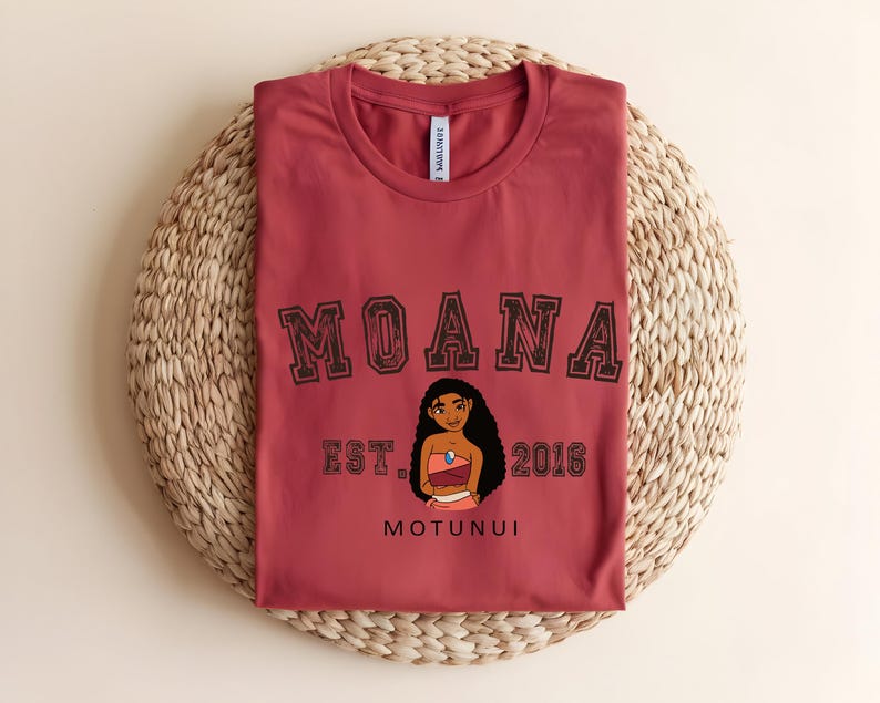 May include: A maroon t-shirt with the word "MOANA" in a collegiate font above a cartoon character illustration. Below the illustration are the words "EST. 2016" and "MOTUNUI". The shirt is folded on a woven, circular surface.