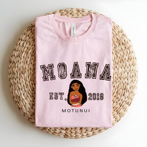 May include: A pale pink t-shirt with the word "MOANA" in a collegiate font above a cartoon character illustration. Below the illustration are the words "EST. 2016" and "MOTUNUI". The shirt is folded on a woven, circular mat.
