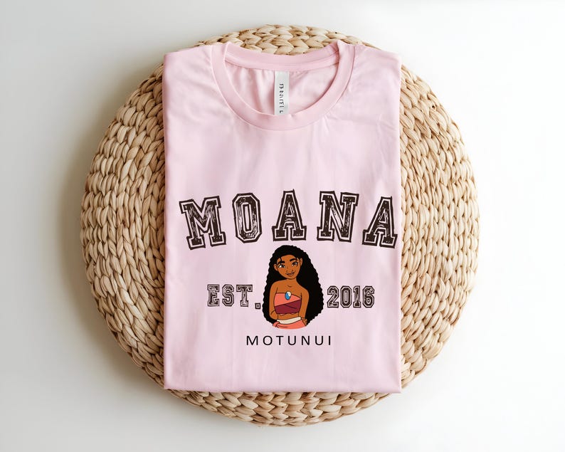 May include: A pale pink t-shirt with the word "MOANA" in a collegiate font above a cartoon character illustration. Below the illustration are the words "EST. 2016" and "MOTUNUI". The shirt is folded on a woven, circular mat.