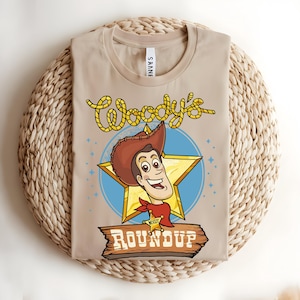 Puede incluir: Camiseta beige con una ilustraci&oacute;n de dibujos animados de Woody de Toy Story. El dise&ntilde;o incluye la cara de Woody, una estrella y el texto "Woody's Roundup". La camiseta est&aacute; doblada sobre una superficie tejida.