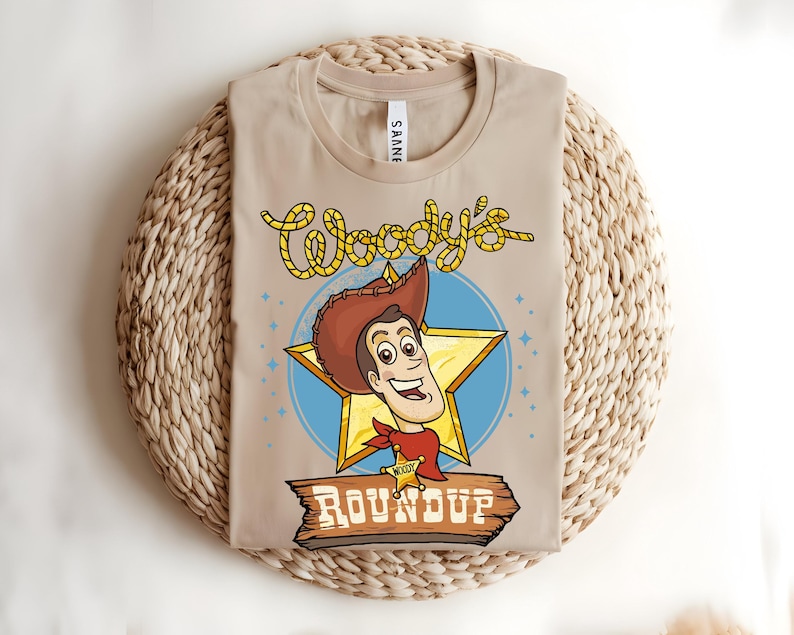 Puede incluir: Camiseta beige con una ilustraci&oacute;n de dibujos animados de Woody de Toy Story. El dise&ntilde;o incluye la cara de Woody, una estrella y el texto "Woody's Roundup". La camiseta est&aacute; doblada sobre una superficie tejida.