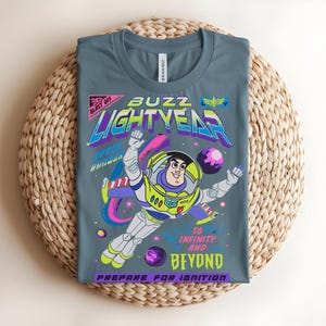 May include: A grey t-shirt with a graphic of Buzz Lightyear in a space scene. The design includes the text "BUZZ LIGHTYEAR" in neon colours, along with the phrases "TO INFINITY AND BEYOND" and "PREPARE FOR IGNITION."