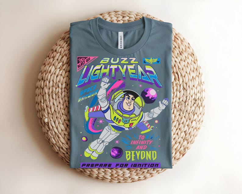 May include: A grey t-shirt with a graphic of Buzz Lightyear in a space scene. The design includes the text "BUZZ LIGHTYEAR" in neon colours, along with the phrases "TO INFINITY AND BEYOND" and "PREPARE FOR IGNITION."