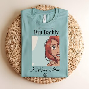 May include: A light blue crew neck t-shirt featuring a graphic of a red-haired character. The text reads EST 1989, But Daddy, and I Love Him, with a small seashell icon at the bottom. The design is centered on the front of the garment.