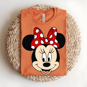 Puede incluir: Una camiseta naranja de cuello redondo con un gran estampado gr&aacute;fico de Minnie Mouse en la parte delantera. El personaje tiene la cabeza negra, la cara de color crema y un lazo rojo brillante con lunares blancos entre dos grandes orejas negras.