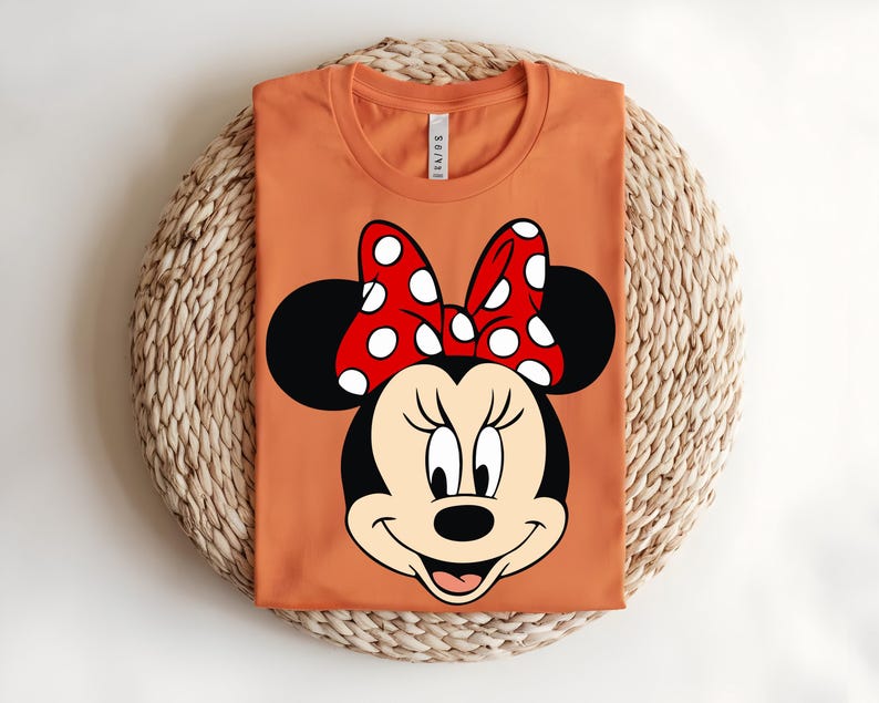 Puede incluir: Una camiseta naranja de cuello redondo con un gran estampado gr&aacute;fico de Minnie Mouse en la parte delantera. El personaje tiene la cabeza negra, la cara de color crema y un lazo rojo brillante con lunares blancos entre dos grandes orejas negras.