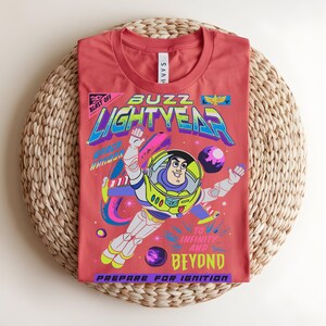 May include: A coral-coloured t-shirt with a graphic of Buzz Lightyear in a space scene. The design includes the words "Buzz Lightyear", "To Infinity and Beyond", and "Prepare for Ignition". The shirt is laid on a woven, circular mat.