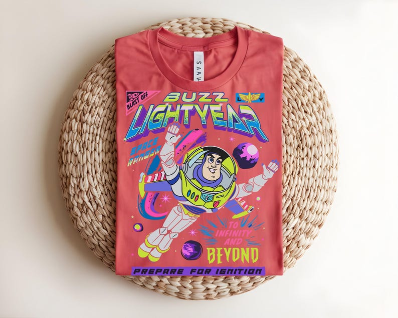 May include: A coral-coloured t-shirt with a graphic of Buzz Lightyear in a space scene. The design includes the words "Buzz Lightyear", "To Infinity and Beyond", and "Prepare for Ignition". The shirt is laid on a woven, circular mat.