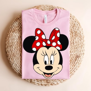 Puede incluir: Una camiseta de color rosa claro con un estampado gr&aacute;fico de la cara de un rat&oacute;n de dibujos animados con grandes orejas negras y un lazo rojo con lunares blancos. El personaje tiene la cara color crema, nariz negra y una sonrisa.