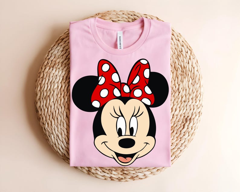 Puede incluir: Una camiseta de color rosa claro con un estampado gr&aacute;fico de la cara de un rat&oacute;n de dibujos animados con grandes orejas negras y un lazo rojo con lunares blancos. El personaje tiene la cara color crema, nariz negra y una sonrisa.