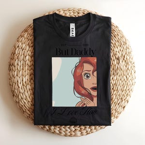 May include: A black t-shirt featuring a graphic print of a red-haired character with wide eyes. The text above reads EST 1989 But Daddy and the text below reads I Love Him, with a small seashell icon centered at the bottom of the graphic design.