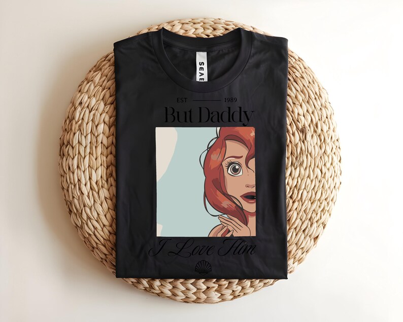 May include: A black t-shirt featuring a graphic print of a red-haired character with wide eyes. The text above reads EST 1989 But Daddy and the text below reads I Love Him, with a small seashell icon centered at the bottom of the graphic design.