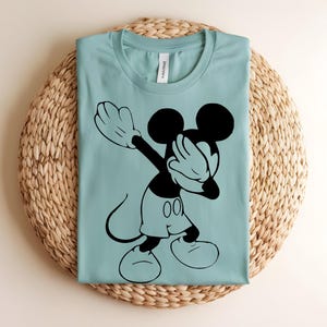 K&ouml;nnte beinhalten: Ein hellblaues T-Shirt mit einem schwarzen Grafikdruck von Mickey Mouse, der eine Dab-Pose ausf&uuml;hrt. Das T-Shirt hat einen Rundhalsausschnitt und ein wei&szlig;es Innenetikett am Nacken. Das Design zeigt die Silhouette der Figur mit Hose.