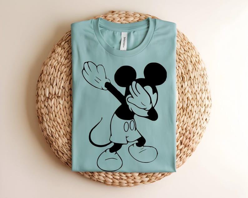 K&ouml;nnte beinhalten: Ein hellblaues T-Shirt mit einem schwarzen Grafikdruck von Mickey Mouse, der eine Dab-Pose ausf&uuml;hrt. Das T-Shirt hat einen Rundhalsausschnitt und ein wei&szlig;es Innenetikett am Nacken. Das Design zeigt die Silhouette der Figur mit Hose.